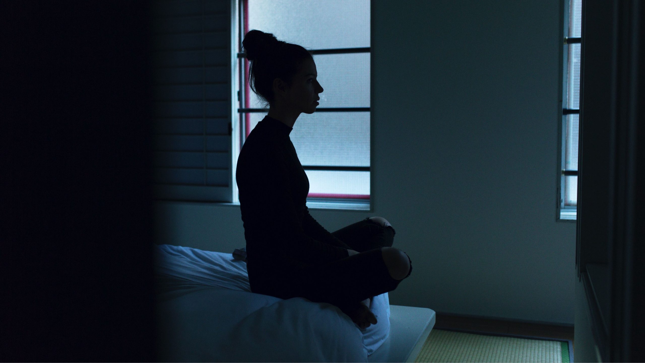 "Depressed woman sitting on the edge of a bed, looking out a window"